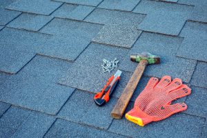 Picture of nails, a cutting tool, a hammer, and a glove on an asphalt shingle roof.